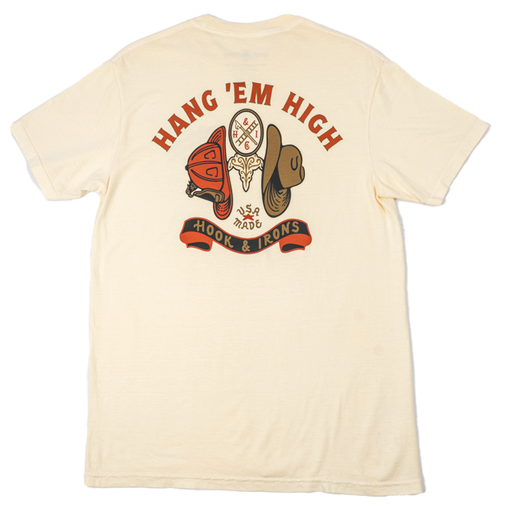 A cream-colored short-sleeve t-shirt shown from the back, featuring a large graphic print with the words 'HANG 'EM HIGH' in red arched lettering above a dreamcatcher, boots, and a banner reading 'Hook & Irons'.