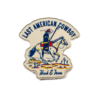 A small enamel pin featuring a firefighter on horseback with a flag and the text 'LAST AMERICAN COWBOY' at the top and 'Hook and Irons' at the bottom, shown on a plain white background.